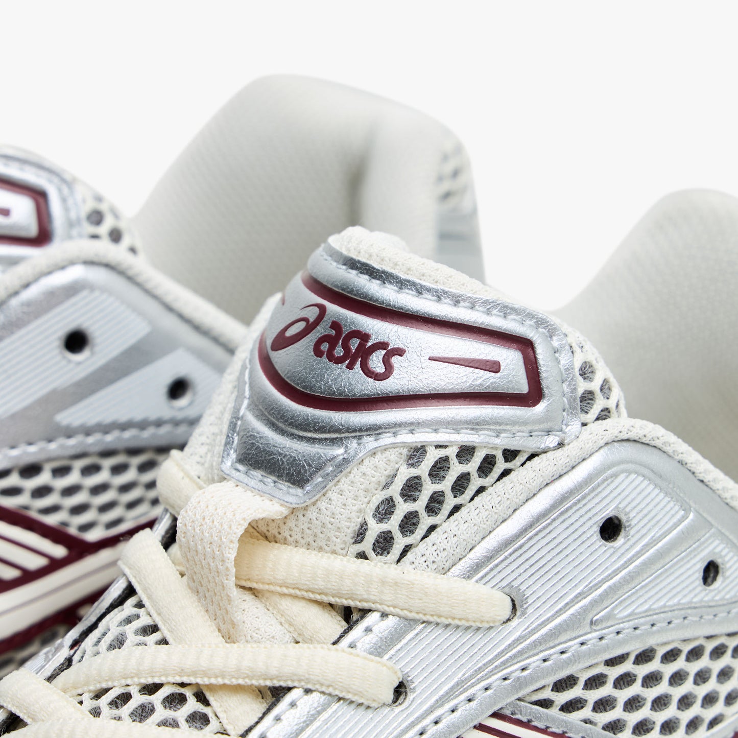 ASICS GEL-KAYANO 14. Close-up of silver overlay on mesh tongue showing red ASICS logo, silver synthetic overlays, and cream laces. 
