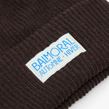 Balmoral Orchard Cotton Beanie. Close-up view highlighting the fine coffee brown ribbed cotton knit construction and the white woven patch sewn onto the cuff with bright blue "BALMORAL AUTOMNE - HIVER" branding. 