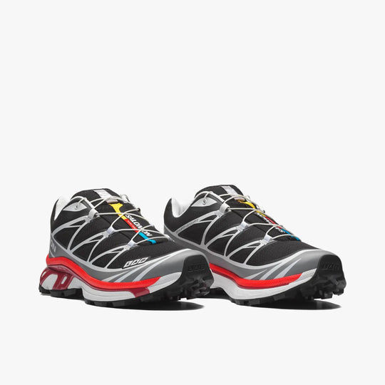 Salomon XT-6. Three-quarter view of a pair of technical trail sneakers displaying black mesh uppers, alloy grey TPU overlays, Quicklace speed lacing system with colorful tongue label accents, lava falls red ACS midsoles, and black outsoles. 