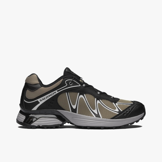 Salomon XT-WHISPER. Side view of trail-inspired lifestyle sneakers featuring a silver sage textile upper, black TPU Sensifit overlays, silver overlay piping, Salomon branding along the collar overlay, black protective toe cap, grey midsole with Agile Chassis System stabilization, and a black lugged rubber outsole. 