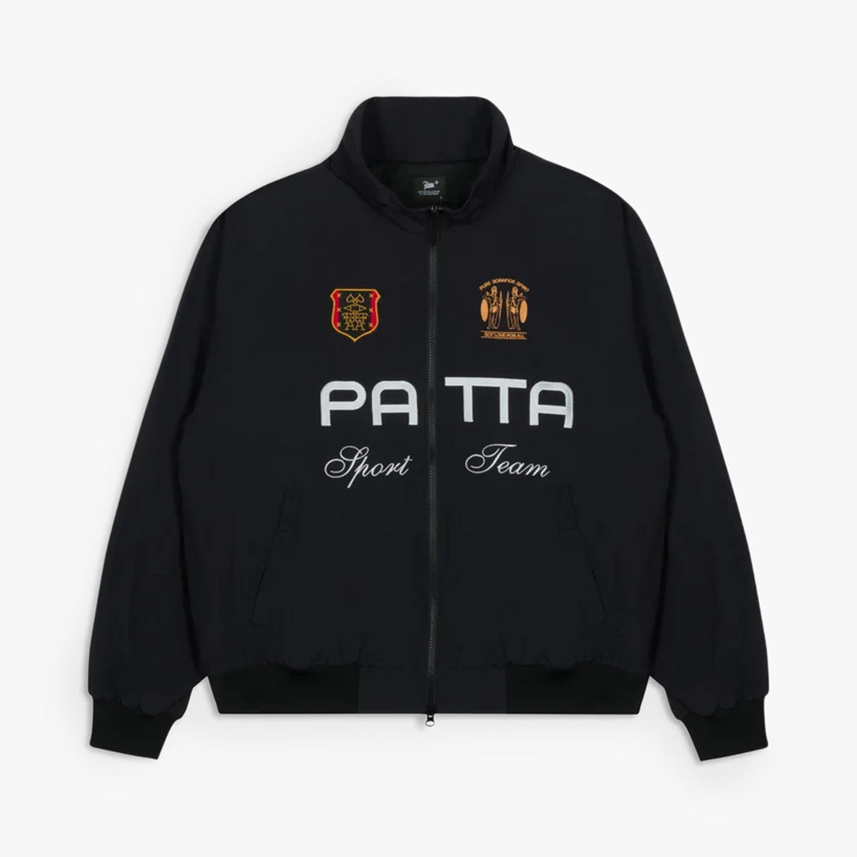 Patta Racing Team Track Jacket. Flat lay front view of a black full-zip track jacket featuring a high stand collar, white printed "PATTA" chest branding with cursive "Sport Team" script underneath, yellow and orange embroidered graphic crests on both sides of the upper chest, full front zip closure, side welt pockets, ribbed cuffs, and a ribbed hem. 