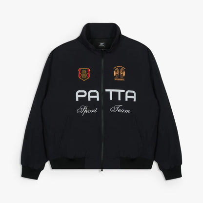 Patta Racing Team Track Jacket. Flat lay front view of a black full-zip track jacket featuring a high stand collar, white printed "PATTA" chest branding with cursive "Sport Team" script underneath, yellow and orange embroidered graphic crests on both sides of the upper chest, full front zip closure, side welt pockets, ribbed cuffs, and a ribbed hem. 