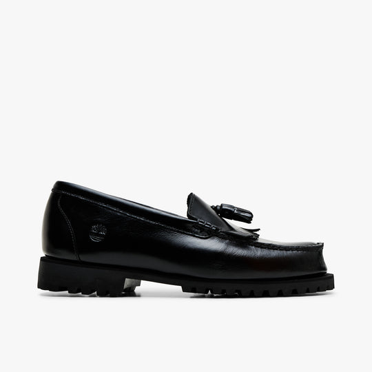 Timberland Authentic Loafer. Lateral profile view featuring polished black full-grain leather upper, kiltie fringe, double tassels, traditional moc-toe stitching, embossed Timberland tree logo on the heel, and rugged lug sole. 