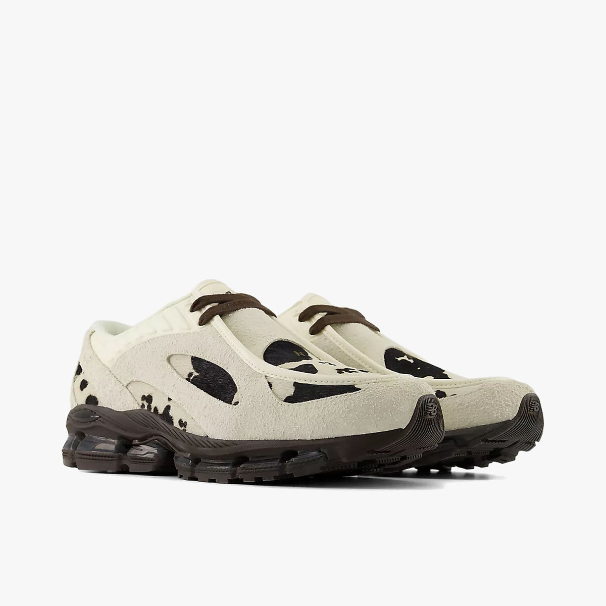 New Balance 1890A Moc-Toe. Three-quarter view of a pair of hybrid moc-toe sneakers displaying angora off-white uppers with dark cow-print graphic overlays, rich earth brown laces, and segmented gel-cushioned midsoles. 
