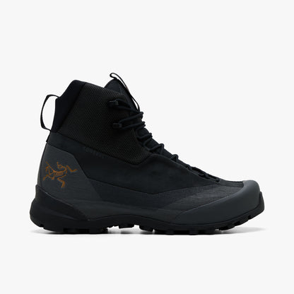 Arc'teryx Konseal Trek. Side view of mid-cut technical hiking boots featuring a black abrasion-resistant textile collar, nubuck upper, dark graphite TPU overlays, gold Archaeopteryx bird logo printed on the lateral heel, webbed lacing loops with speed hooks, and a lugged black Vibram outsole. 