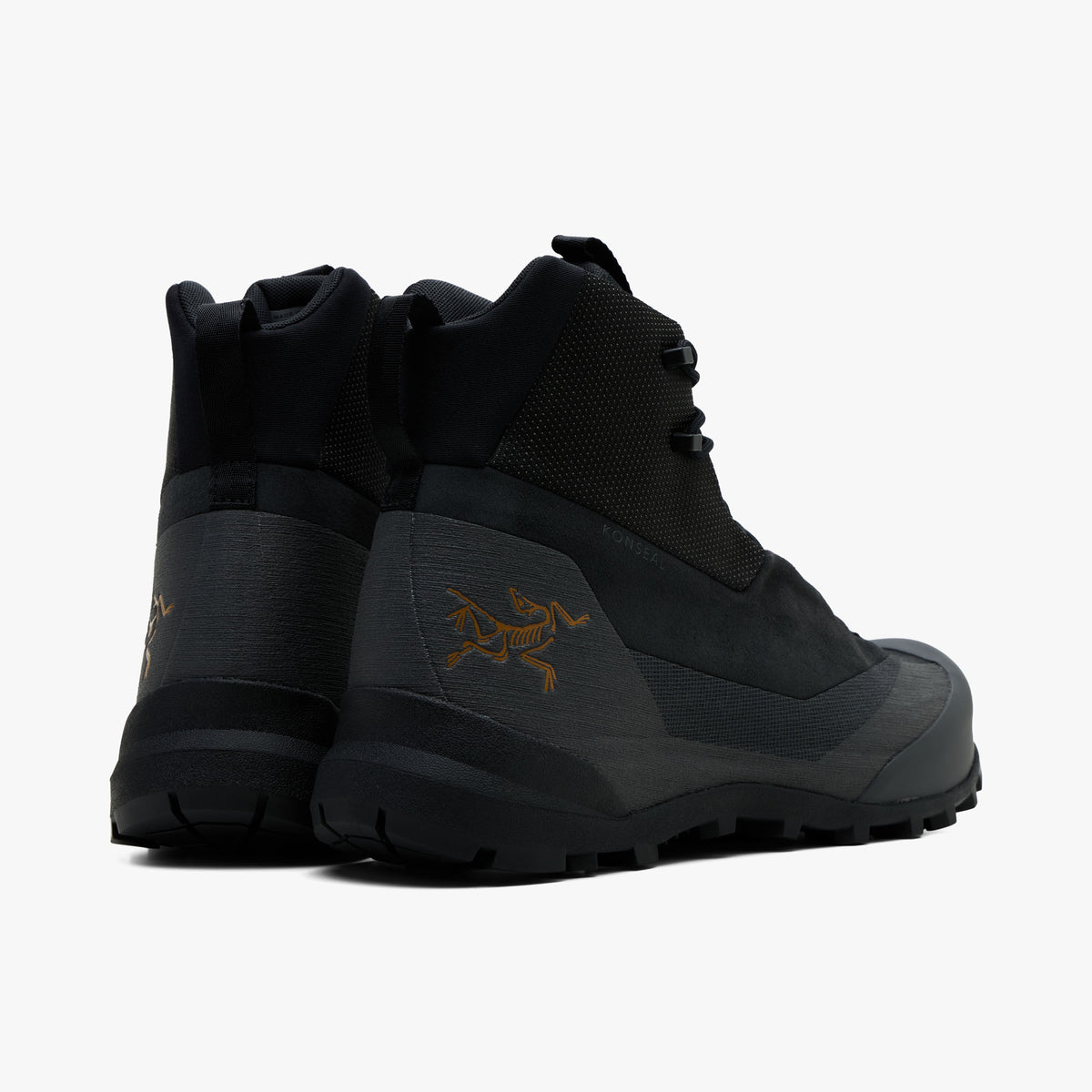 Arc'teryx Konseal Trek. Rear view of a pair of hiking boots showing black high-top collar shafts, dark graphite synthetic heel counters with gold Archaeopteryx bird logos, heel pull loops, and black midsoles. 