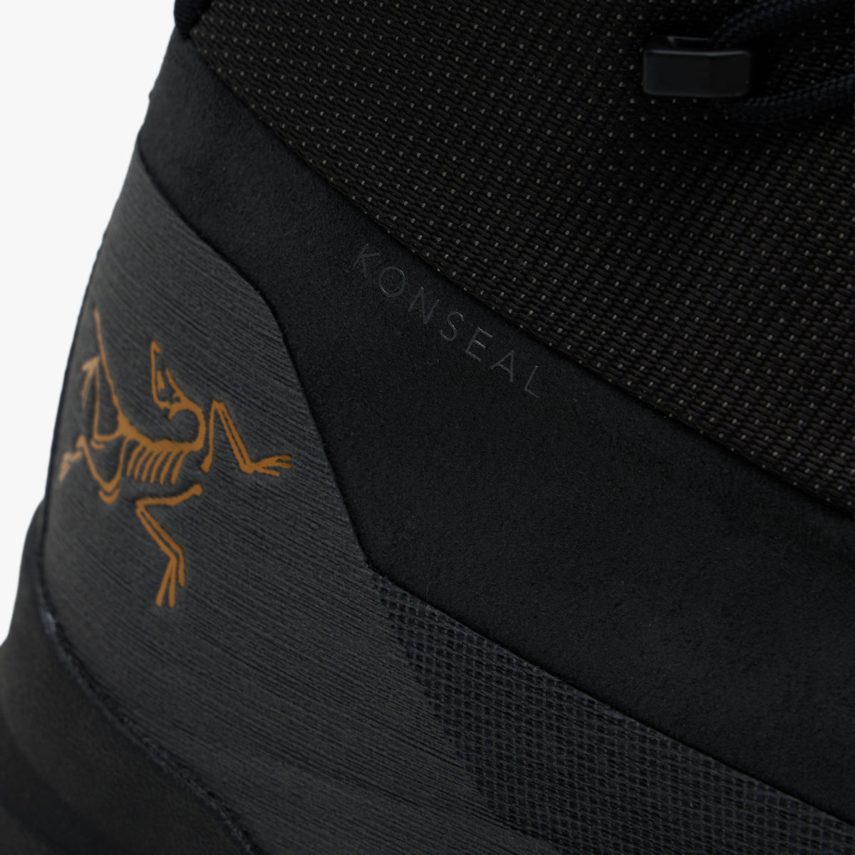 Arc'teryx Konseal Trek. Close-up lateral rear angle displaying dark graphite synthetic heel counter, subtle KONSEAL model branding, gold printed Archaeopteryx bird logo, and black foam midsole edge. 